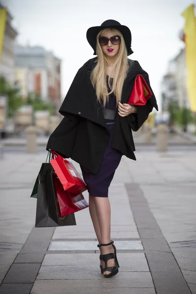 Linda jovem mulher loira elegante em vestido azul, casaco, óculos de sol e chapéu com sacos de compras andando na cidade — Fotografia de Stock