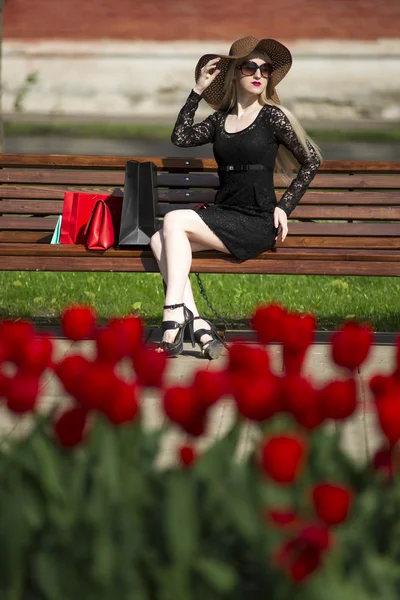 Όμορφη νεαρή κομψό ξανθιά γυναίκα στο μαύρο φόρεμα, γυαλιά ηλίου και το καπέλο με τσάντες αγορών που κάθεται σε ένα παγκάκι στο πάρκο — Φωτογραφία Αρχείου