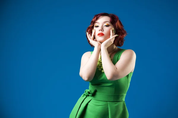 Young beautiful red-haired caucasian woman in green dress posing in studio on blue background, professional makeup and hairstyle — Stock Photo, Image