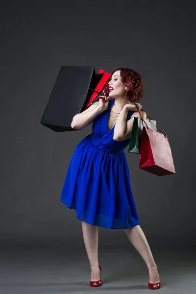 Conceito Shopaholic, jovem bela mulher caucasiana ruiva em vestido azul posando em estúdio em fundo cinza, maquiagem profissional e penteado — Fotografia de Stock