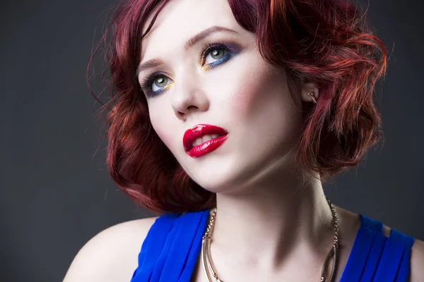 Young beautiful red-haired caucasian woman in blue dress posing in studio on gray background, professional makeup and hairstyle — Stock Photo, Image