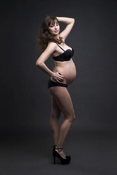 Jeune beauté et sexy femme enceinte caucasienne en lingerie noire et chaussures à talons hauts sur fond de studio gris — Photo