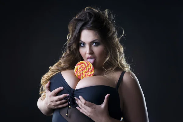 Giovane bella caucasico plus size modello con grande seno in reggiseno nero leccare un lecca-lecca, xxl donna su sfondo scuro — Foto Stock