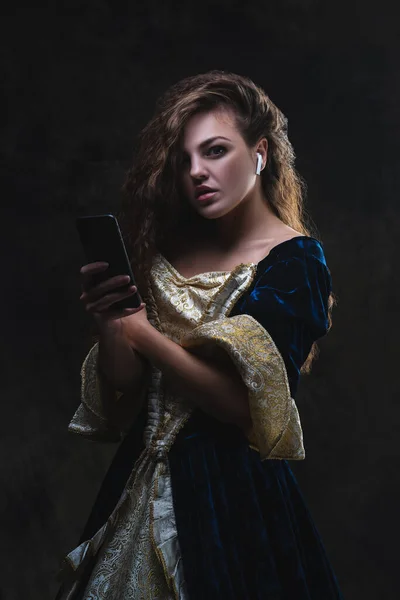 Beautiful woman in renaissance dress listens to music in wireless headphones on abstract dark background, old and new concept