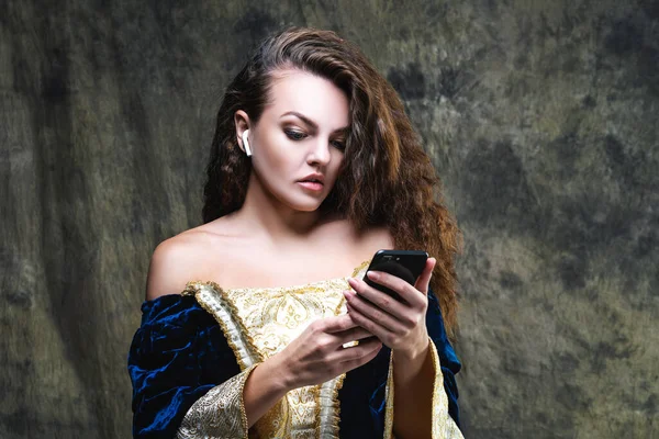 Beautiful woman in renaissance dress listens to music in wireless headphones on abstract dark background, old and new concept