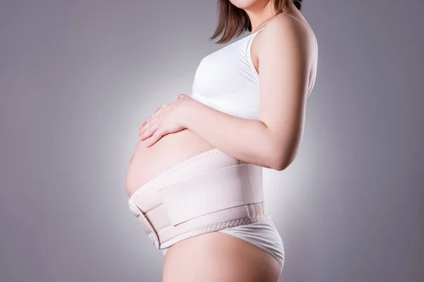 Pregnant woman in white underwear with orthopedic support belt, pregnancy bandage, studio shot on gray background