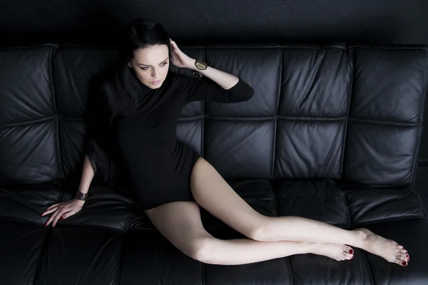 Красивая молодая женщина в телесном костюме с длинными ногами и длинными черными волосами сидит на черном диване — стоковое фото