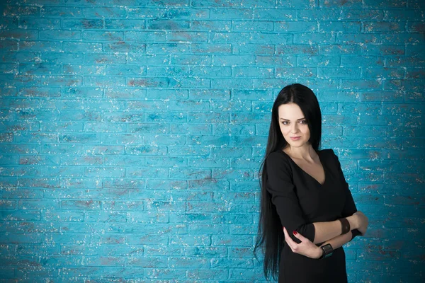 Schöne junge Frau in einem schwarzen Kleid posiert vor dem Hintergrund einer blauen Backsteinmauer — Stockfoto