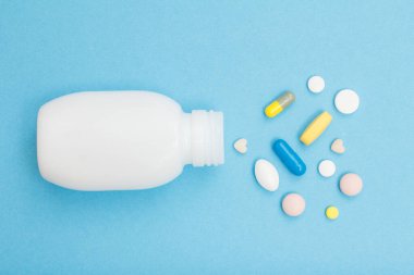Mavi arka planda kalp şeklinde haplar ve kapsüllerle dolu beyaz bir şişe. Üst görünüm