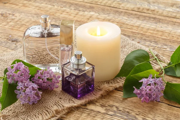 Bottles of perfume, a burning candle and lilac flowers on sackcloth and old wooden boards.