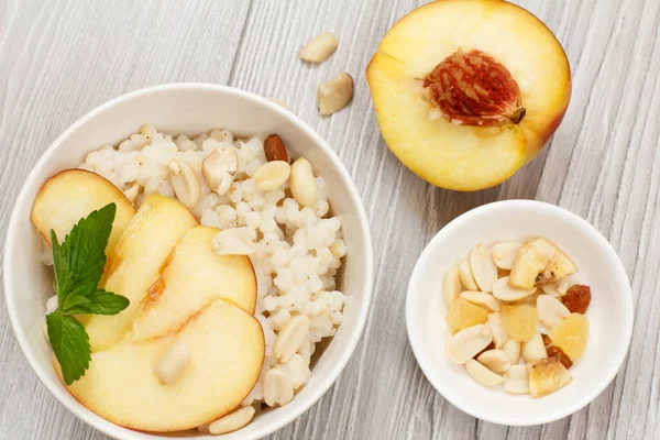 Sorghum porridge with pieces of peach, cashew nuts and almond in porcelain bowls, a fresh peach on the gray wooden boards. Vegan gluten-free sorghum salad with fruits.