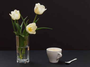 Cam vazoda sarı lale demeti ve siyah arka planda kaşık olan bir fincan kahve..