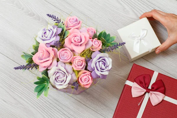 Woman's hand holding a gift box over gray wooden background with soap in form of roses. Concept of giving a gift on holidays. Top view.
