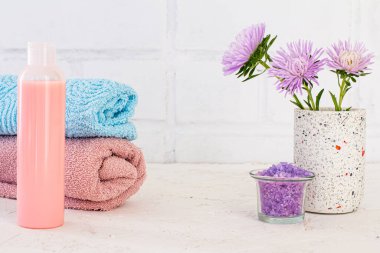 Deniz tuzu, bir şişe şampuan, havlular ve beyaz arka planda aster çiçekleri. Kadın kozmetik ürünleri ve aksesuarları.