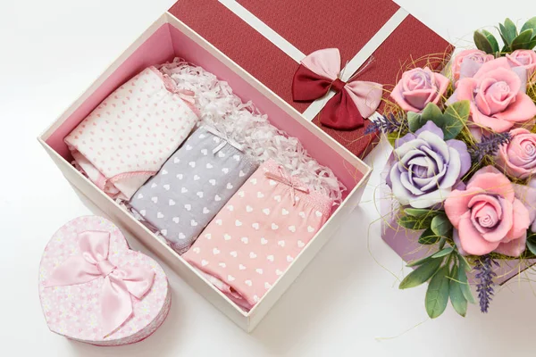 Beyaz arka planda suni çiçekler olan bir kutuda farklı renklerde katlanmış pamuk külotlar. Kadın iç çamaşırı seti. Üst görünüm.