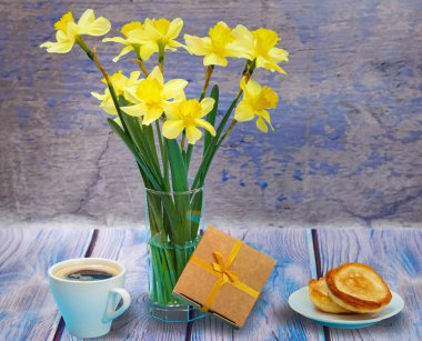 Cam vazoda bir buket sarı nergis, bir hediye kutusu, bir fincan kahve ve ahşap zemin üzerinde krep olan bir tabak. Boyalı resim efekti.