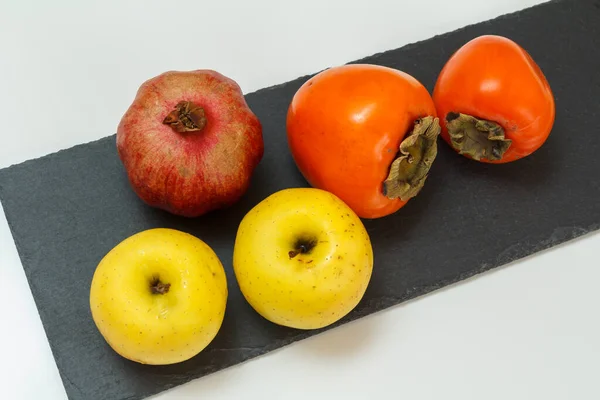 Apples, pomegranate and ripe persimmon fruit on the black stone board. — Stock Photo, Image