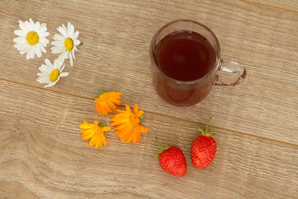 Glass cup of tea with white chamomile flowers, yellow marigold flowers and strawberries on the wooden desktop. Top view.