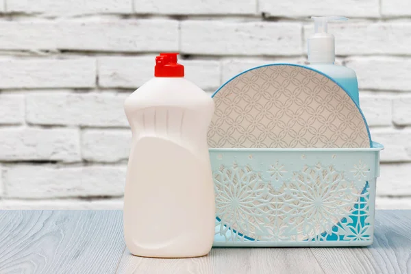 Plastic bottles of dishwashing liquid, glass and tile cleaner, a clean plate in a basket with a white wall on the background. Washing and cleaning concept.