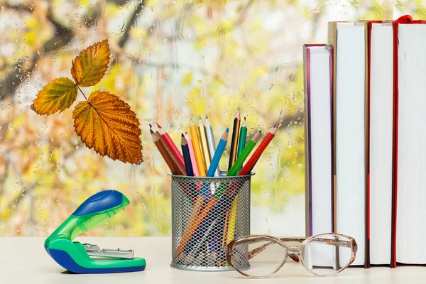 Books, colour pencils, glasses, stapler on desk in front of the window with water drops.