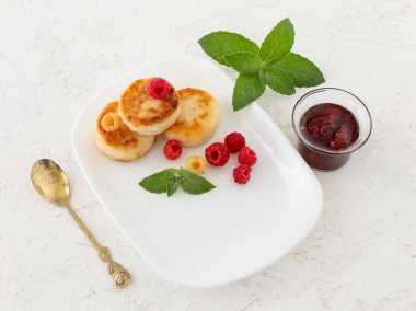 White porcelain plate with homemade curd fritters, raspberries and mint leaves, a glass bowl with strawberry jam, a teaspoon on the white background. Top view.