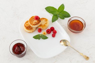 White porcelain plate with homemade curd fritters, raspberries and mint leaves, a teaspoon, glass bowls with strawberry jam and honey on the white background. Top view.