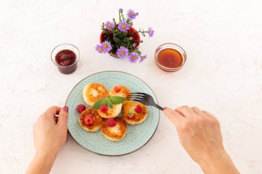 Çatallı bir kadın elleri ve ev yapımı kremalı böreklerle süslenmiş ahududu ve nane yapraklı bir tabak. Çilek reçelli cam kaseler ve bal, beyaz arka planda çiçekli vazo. Üst görünüm.