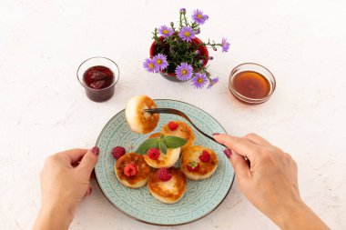 Çatallı bir kadın elleri ve ev yapımı kremalı böreklerle süslenmiş ahududu ve nane yapraklı bir tabak. Çilek reçelli cam kaseler ve bal, beyaz arka planda çiçekli vazo. Üst görünüm.