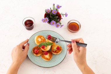 Çatallı bir kadın elleri ve ev yapımı kremalı böreklerle süslenmiş ahududu ve nane yapraklı bir tabak. Çilek reçelli cam kaseler ve bal, beyaz arka planda çiçekli vazo. Üst görünüm.
