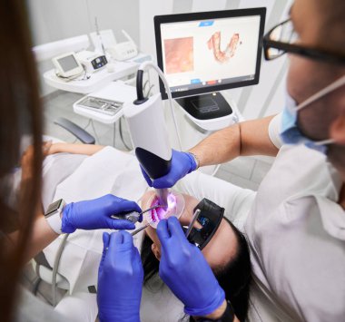 Diş hekimi ve asistanı, diş muayenehanesinde hasta dişlerini incelerken intraoral tarayıcı kullanıyor. Diş uzmanı modern tarama makinesiyle intraoral tarama yapıyor. Dijital dişçilik kavramı