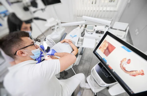 Tıbbi maskeli dişçi ve asistan diş hekiminde dişçi dişlerini incelerken diş içi tarayıcı kullanıyor. Modern tarama makinesi ile intraoral tarama. Dijital dişçilik kavramı.