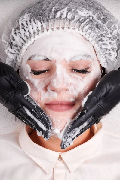 美容师双手紧握黑色无菌手套 在病人脸上涂上清洁泡沫 美容院实施护肤程序 头戴一次性浴帽的年轻女子魅力十足 — 图库照片