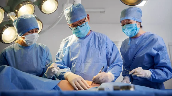 Doctors in blue surgical suits performing aesthetic surgery while patient lying on operating table. Concept of medicine, abdominoplasty and plastic surgery.