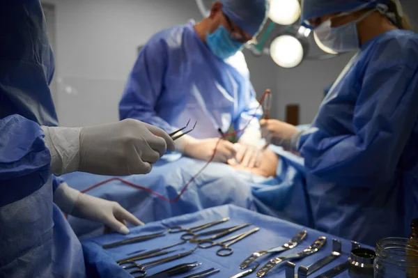 Close up view of assistant\'s hand holding tweezers. On the blurred background surgical team doing plastic surgery in operating room with medical instruments. Concept of medicine and aesthetic surgery.