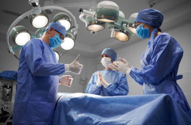 Steril eldivenli ve koruyucu maskeli bir grup doktor başarılı bir ameliyattan sonra hastalarının yanında olacak. Erkek estetik cerrah baş parmak hareketi yaparken asistanlar alkışlıyor.
