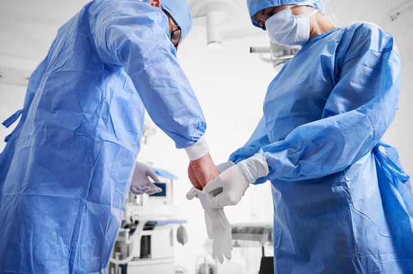 外科医が滅菌手袋を着用するのを助ける青い外科用スーツのアシスタント 診療所で整形手術の準備をしている保護医療用マスクの医師2人 薬や手術の準備の概念 — ストック写真