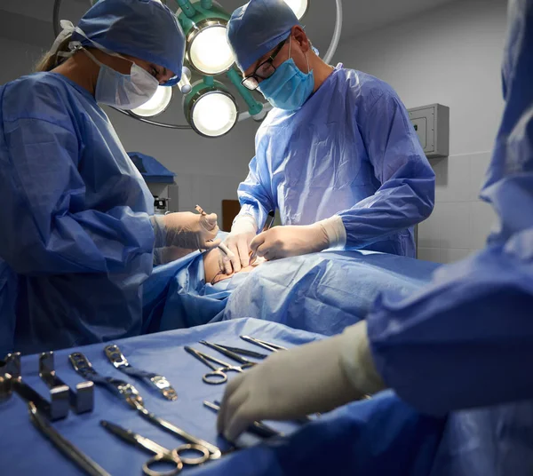 Medical team in surgical suits performing aesthetic surgery in operating room. Doctors doing cosmetic surgery while assistant standing by surgical table with medical tools. Concept of plastic surgery.