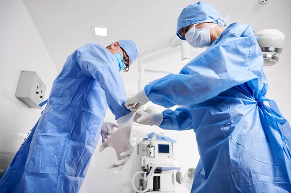 外科医が滅菌手袋を着用するのを助ける青い外科用スーツのアシスタントの側面図 診療所で整形手術の準備をしている保護医療用マスクの医師2人 外科学の概念 — ストック写真