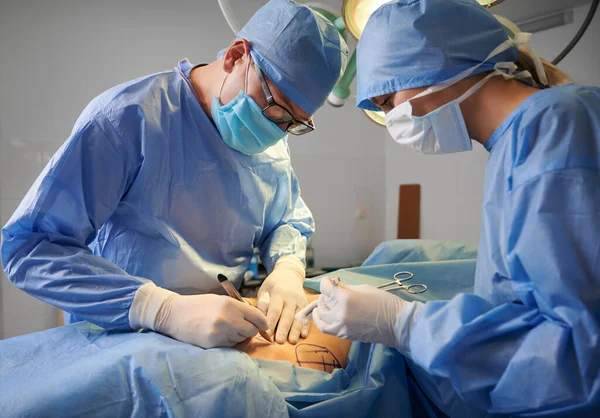 Doctors in surgical hospital suits performing cosmetic surgery in operating room. Medical team wearing protective face masks, sterile gloves and medical caps. Concept of medicine and plastic surgery.