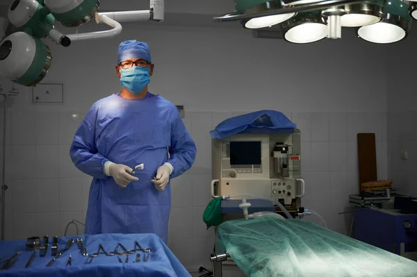 Front view of plastic surgeon holding medical instruments and looking at camera while standing in operating room at hospital. Concept of medicine, medical workers and surgery.