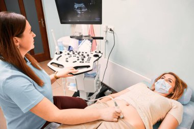 Sonograf, genç bir kadın için ultrason taraması yapıyor. Ultrasonografi sırasında yüz maskesi takan kadın hasta gündüz yatağında yatıyor. Sağlık, tıbbi muayene ve sonografi kavramı.
