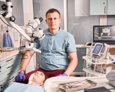 Mavi gömlekli bir erkek dişçinin portresi modern ekipmanlarla dişçi ofisinde genç bir kadının yanında oturuyor. Diş sağlığı, stomatoloji ve diş bakımı kavramı.