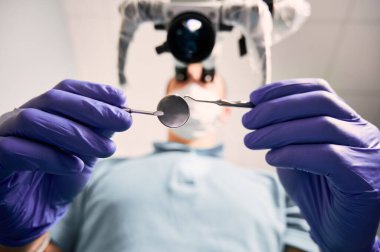 Erkek stomatolojistin, paslanmaz çelik diş kâşifi ve ayna tutan steril eldivenlerini yakın plan. Diş tanılama mikroskobu kullanan erkek dişçi. Diş hekimliği kavramı.