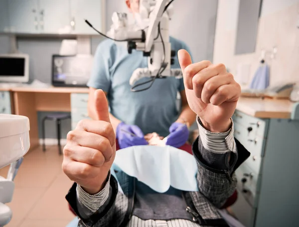 年轻女子躺在牙椅上时 接近她 表示赞同 男牙医检查女病人的牙齿而女病人放弃大拇指 牙科护理和批准的概念 — 图库照片