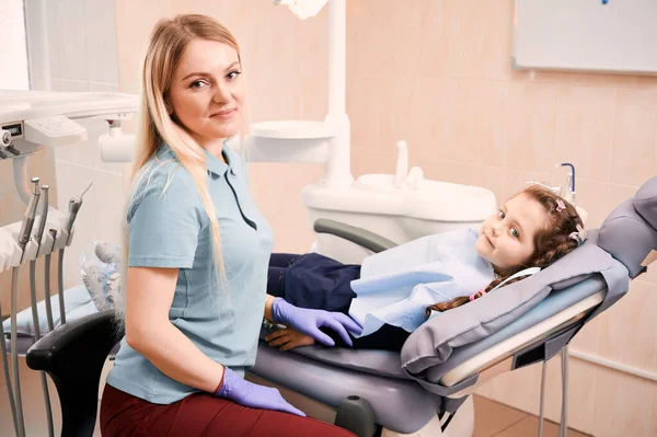 女性の胃科医の横にある歯科椅子に寝そべっている愛らしい子供 歯科事務所で可愛い女の子の横に座っている滅菌手袋の若い女性歯科医 小児科 歯科医療の概念 — ストック写真
