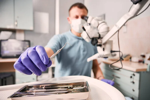 Close up of male dentist hand in sterile glove holding metal dental explorer. Stomatologist in medical mask taking dental instrument while sitting near diagnostic microscope. Concept of dentistry.