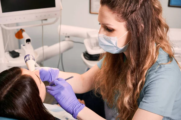 Young female dentist in medical mask and sterile gloves checking patient teeth. Woman lying in dental chair while receiving dental treatment in modern medical center. Concept of dentistry.