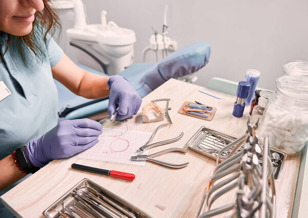Dentist cutting braces wire while sitting at the table with orthodontic instruments. Woman orthodontist preparing wire for braces attachment. Concept of dentistry and orthodontic treatment.