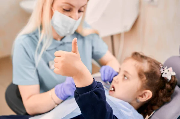子供の横に座って子供の歯を調べる医療マスクの女性の胃科医ながら 女の子の手を親指を与えることに焦点を当てます 小児科 歯科医療承認の概念 — ストック写真