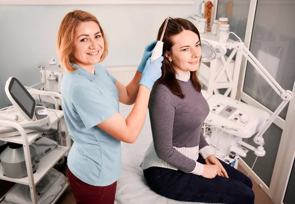 Fröhlicher Arzt Trichologe Bürstet Weibliche Kundenhaare Mit Einem Laser Haarwuchsgerät — Stockfoto